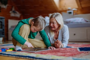 A boy with Down syndrome with his grandmother sitting on floor and using tablet at home.