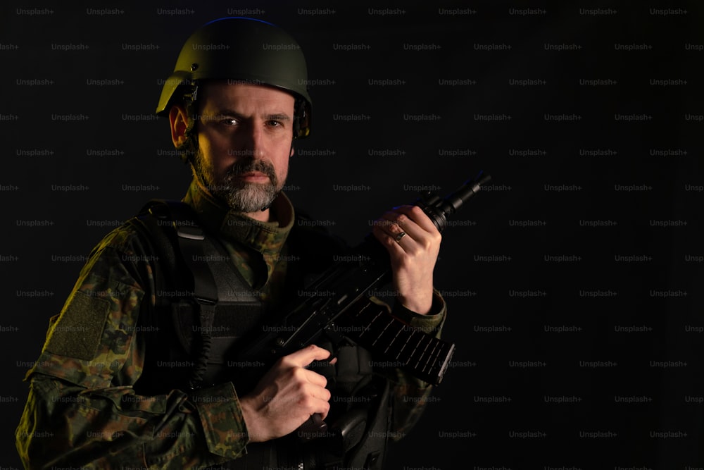 A soldier in military uniform and helmet with weapon looking at camera on black background.