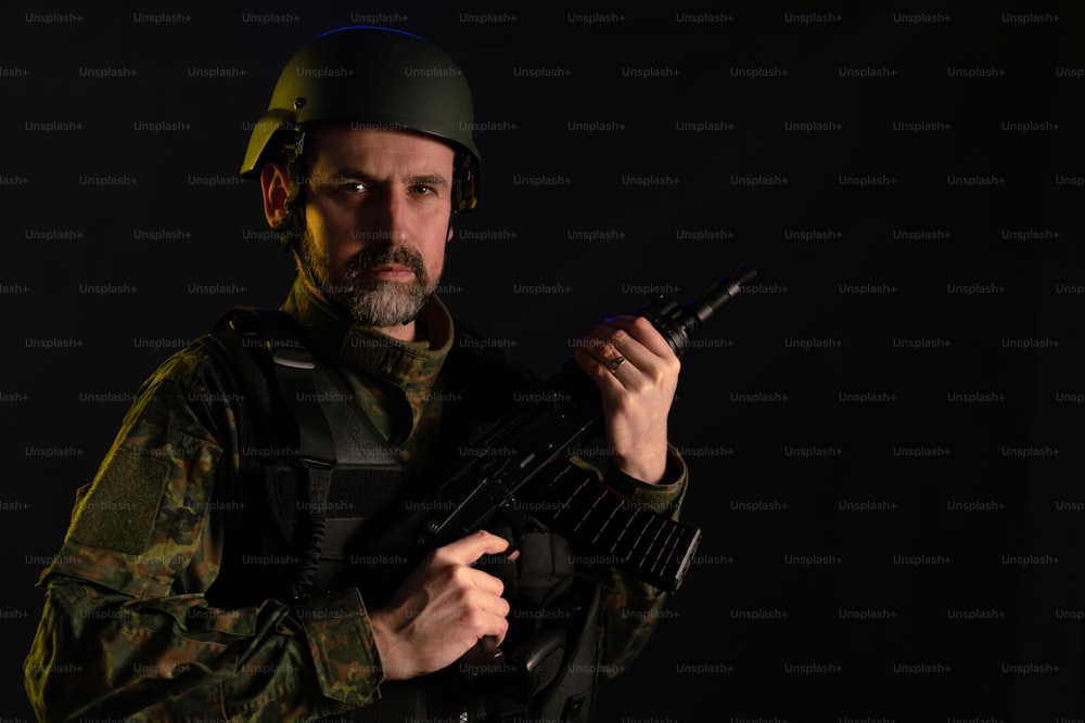 Un soldato in uniforme militare ed elmetto con arma che guarda la macchina fotografica su sfondo nero.