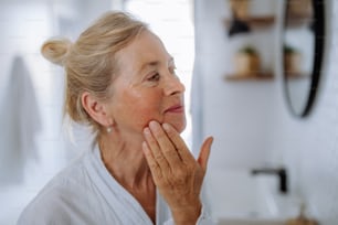 Una bella donna anziana in accappatoio che guarda lo specchio e applica la crema naturale per il viso in bagno, concetto di cura della pelle.