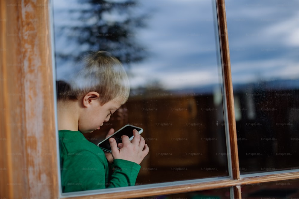 Un niño con síndrome de Down usando un teléfono inteligente en casa, disparado a través de la ventana.
