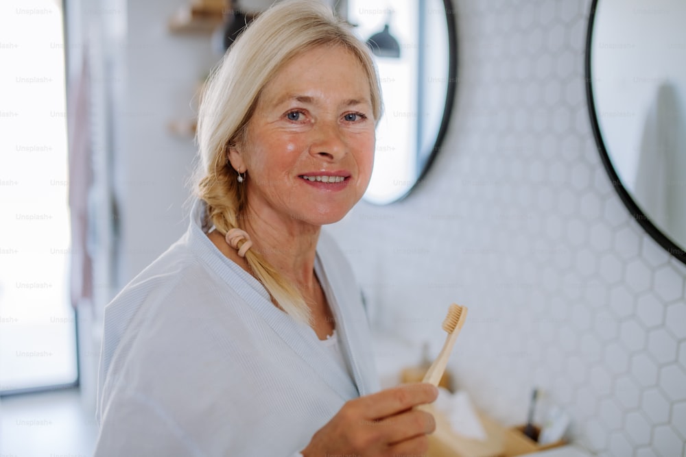 Una hermosa mujer mayor en albornoz cepillarse los dientes con cepillo de dientes de madera ecológica en el baño, estilo de vida sostenible.