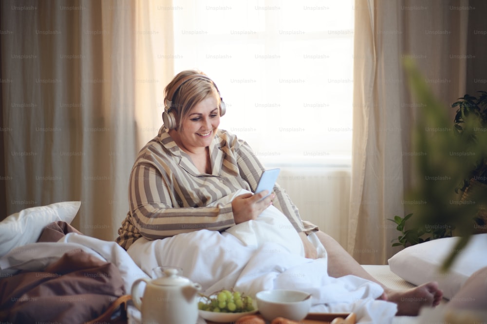 Una felice donna in sovrappeso con cuffie e smartphone che fa colazione a letto a casa.