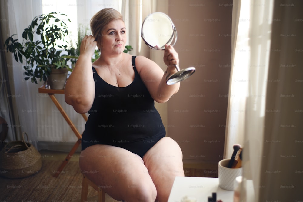 Una donna grassa seduta e guardando lo specchio a casa, concetto di cura di sé.