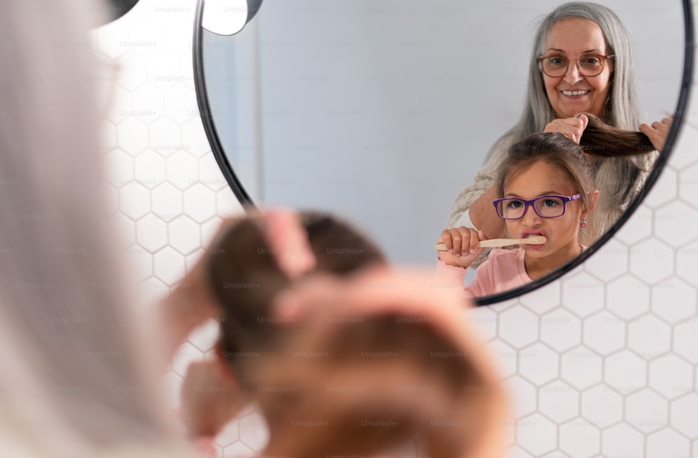 A senior grandmother and granddaughter standing indoors in bathroom, brusing teeth and hair in morning