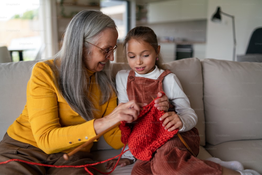 A little girl sitting on sofa with her grandmother and learning to knit indoors at home.
