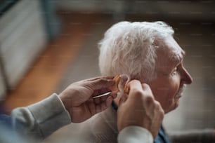 A close-up of caregiver man's hand inserting hearing aid in senior's man ear.