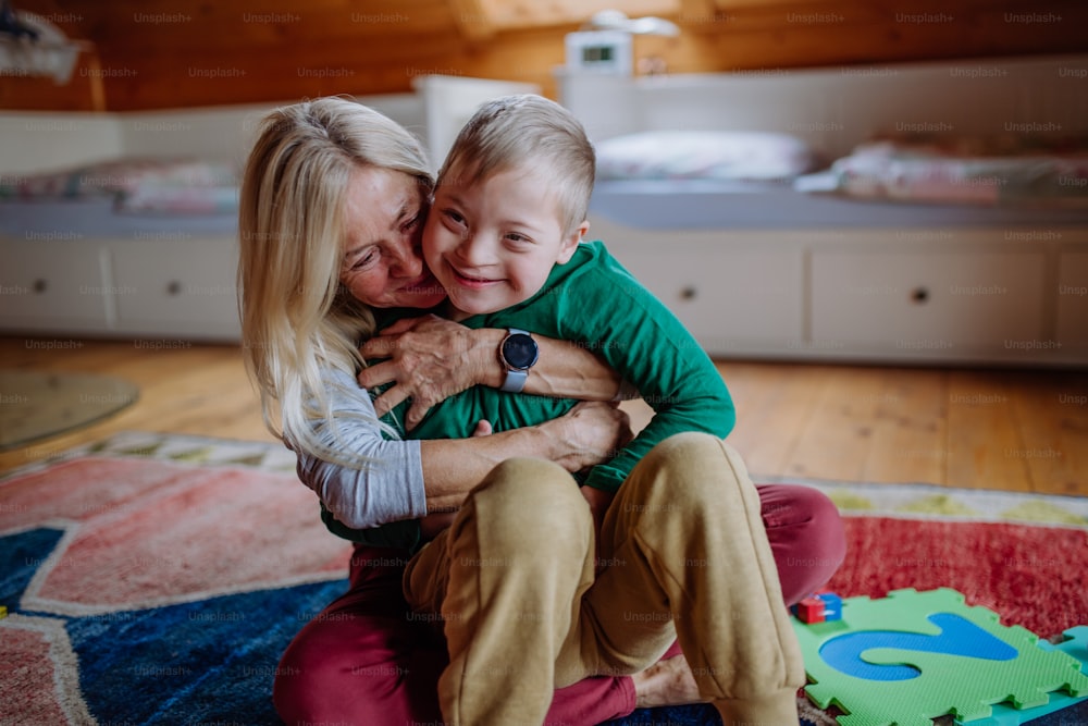 Un niño feliz con síndrome de Down sentado en el suelo y abrazado con su abuela en casa