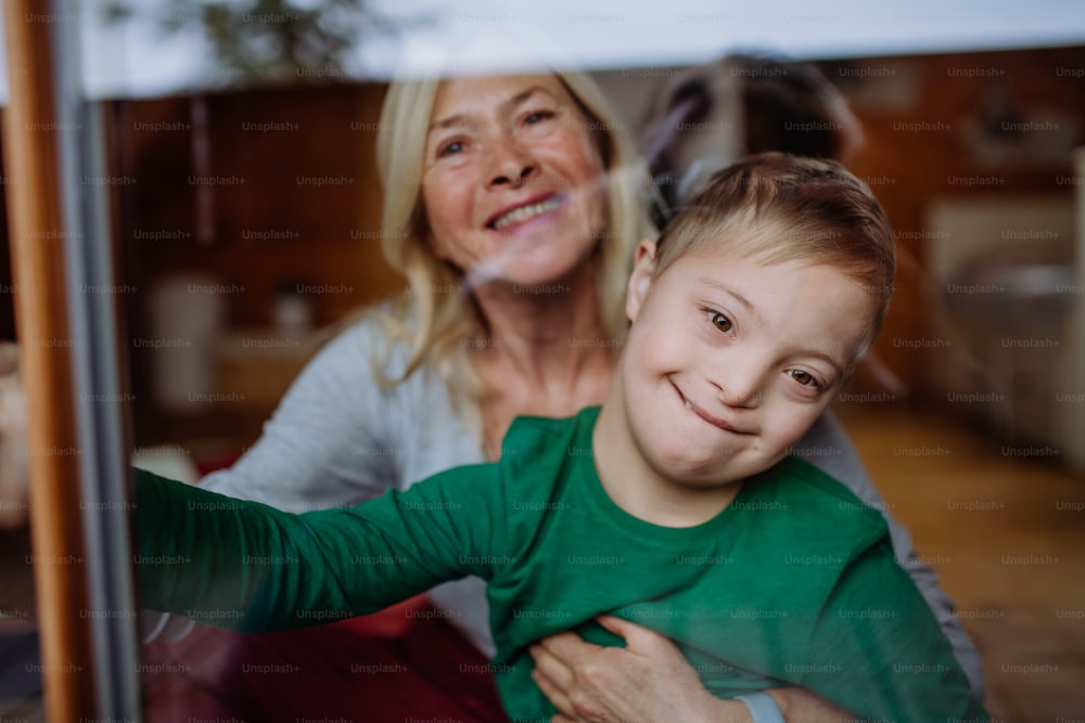 Un niño con síndrome de Down con su abuela mirando a la cámara a través de la ventana de su casa.