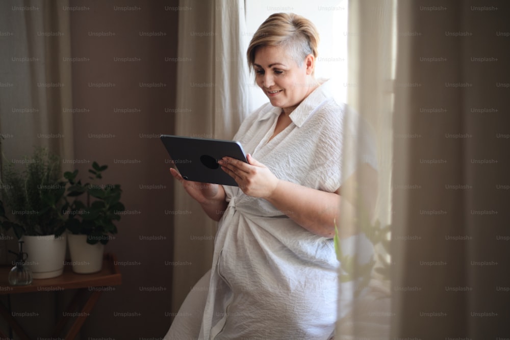 An overweight woman in underwear standing by window and using tablet.