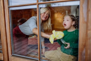 다운 증후군을 앓고있는 소년이 어머니와 할머니와 함께 집에서 창문을 청소하고 있습니다.