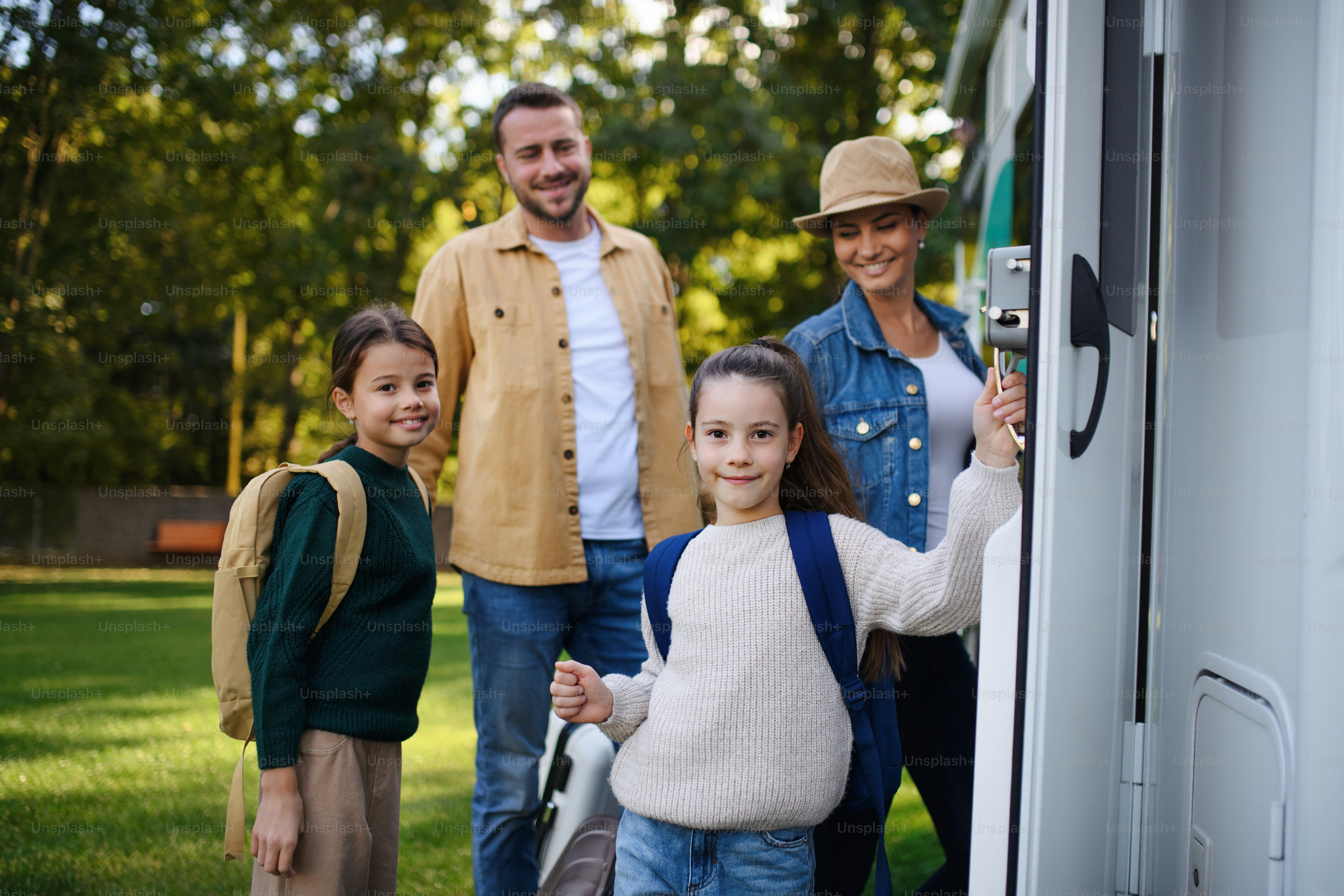Una giovane famiglia felice con i bagagli che guardano la macchina fotografica con il camper sullo sfondo all'aperto in giardino.
