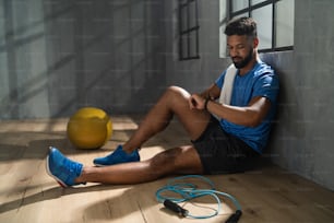 Un joven deportista afroamericano sentado y usando un reloj inteligente en interiores en el gimnasio, concepto de entrenamiento de entrenamiento.