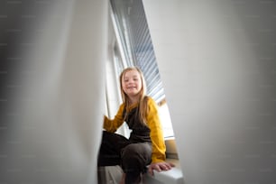 다운 증후군을 앓고있는 행복한 어린 소녀가 집에서 창문에 앉아 있습니다.