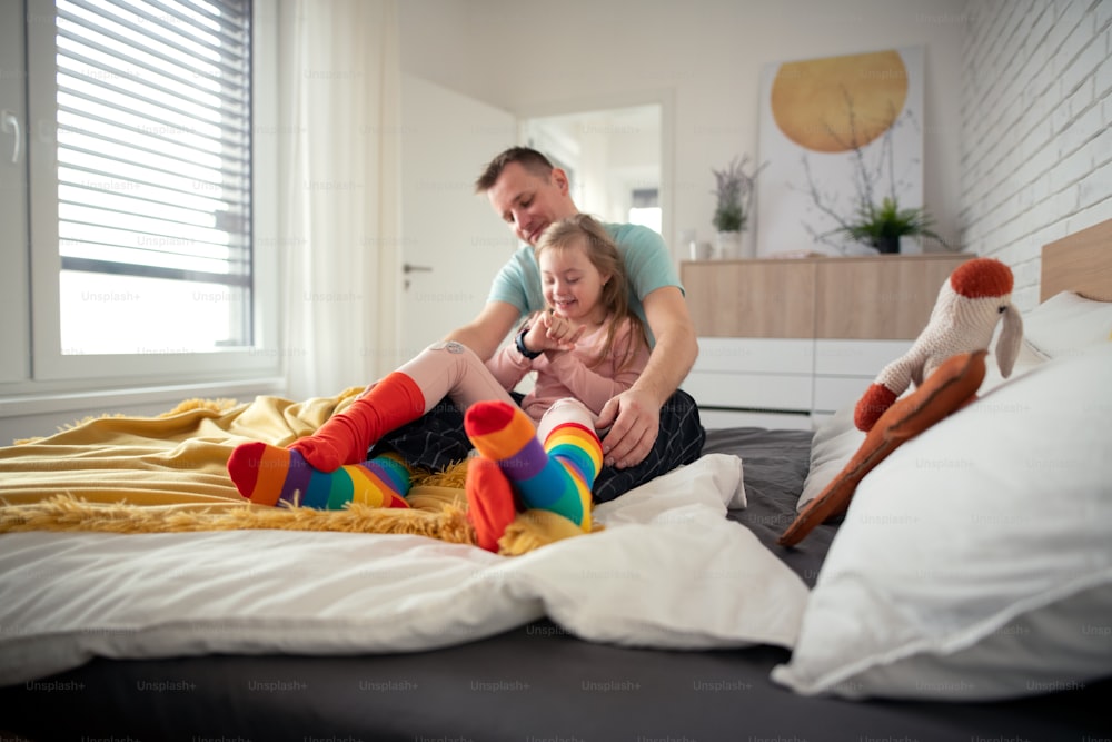 A father putting on different socks to his little daughter with Down syndrome when sitting on bed at home.