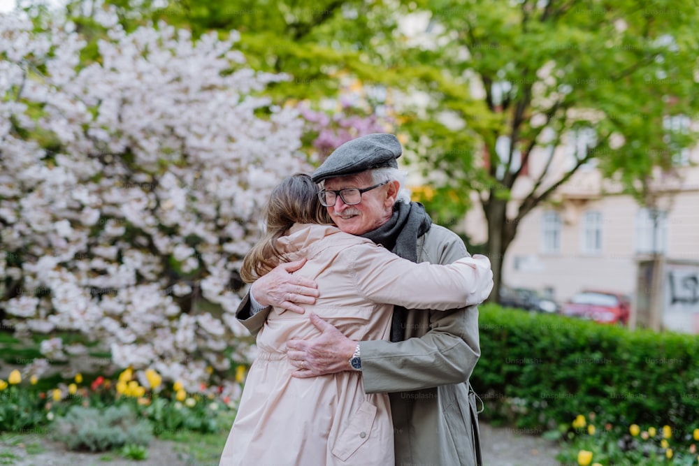An adult daughter hugging her senior father outdoors in park on spring day.