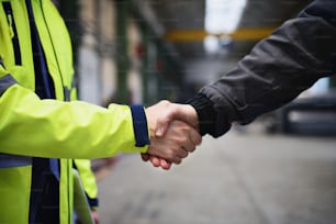 An engineer and industrial worker in uniform shaking hands in large metal factory hall and talking. Close-up.