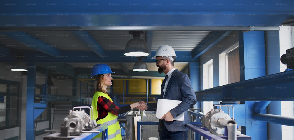 An engineer and industrial worker in uniform shaking hands in large metal factory hall and talking.