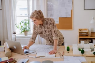 Une femme architecte senior avec une maquette de maisons regardant des plans dans un bureau.