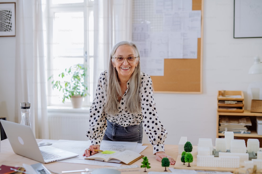 Heureuse femme architecte senior avec un modèle de maisons debout dans un bureau et regardant la caméra.