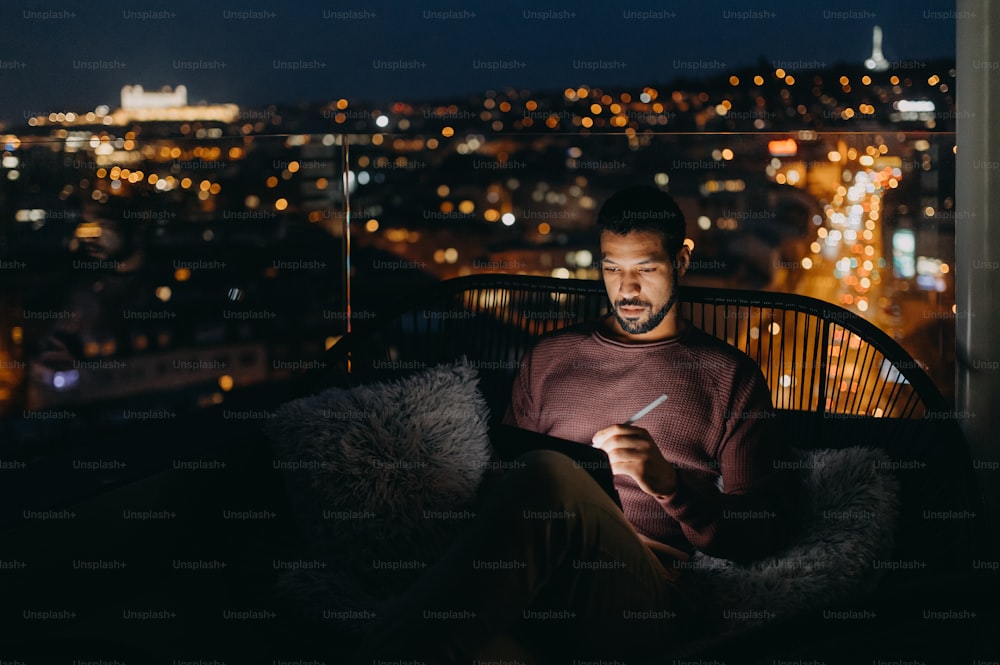 A young African AMerican man sitting on balcony with urban view and using tablet at night