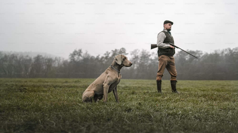 산탄총을 들고 들판에서 전통적인 사격복을 입은 개를 가진 사냥꾼.