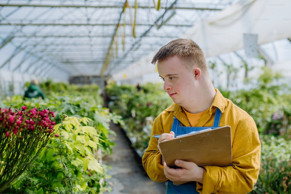 Um jovem com síndrome de Down trabalhando em um centro de jardinagem, holidng prancheta e verificando plantas.