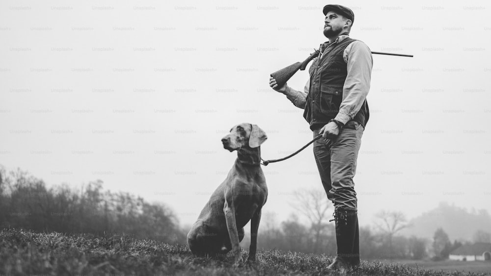 Un uomo cacciatore con cane in abiti da tiro tradizionali sul campo che tiene il fucile, foto in bianco e nero.