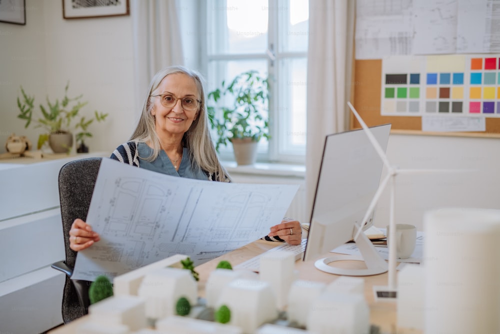 Une éco-architecte senior avec des plans travaillant sur lapotp dans son bureau.