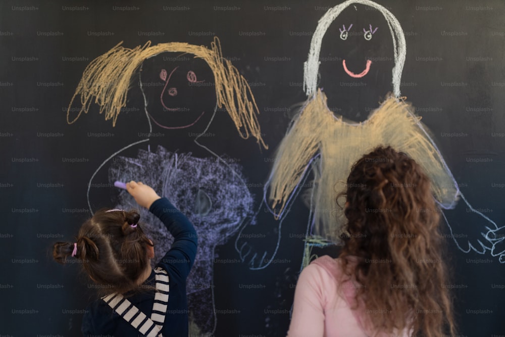 プレイルームの室内で黒板の壁にチョークで絵を描いている小さな女の子のグループの後ろ姿。