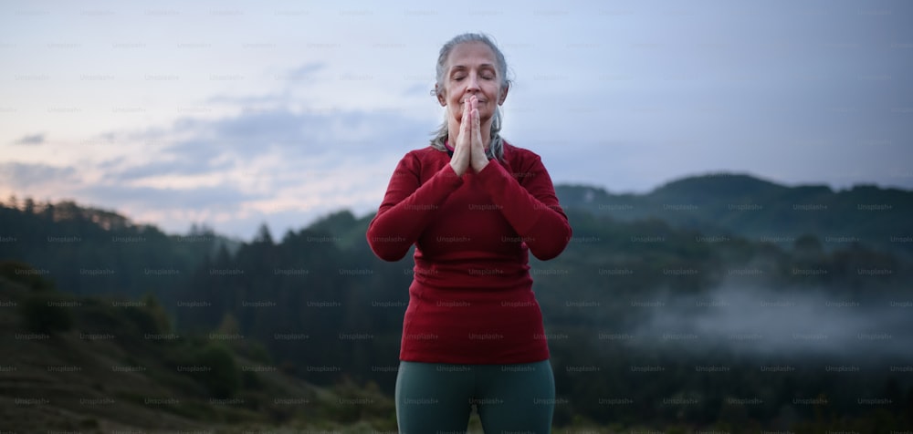 Uma mulher idosa fazendo exercícios de respiração na natureza no início da manhã com neblina e montanhas ao fundo.