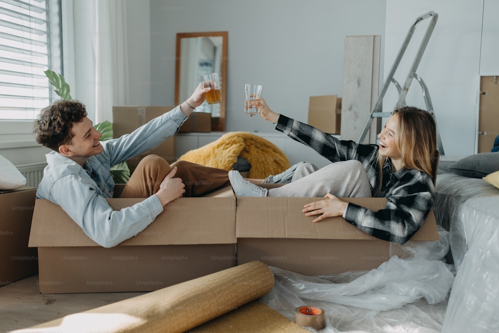 Una alegre pareja joven en su nuevo apartamento, sentados en cajas y tintineando vasos. Concepción de la mudanza.