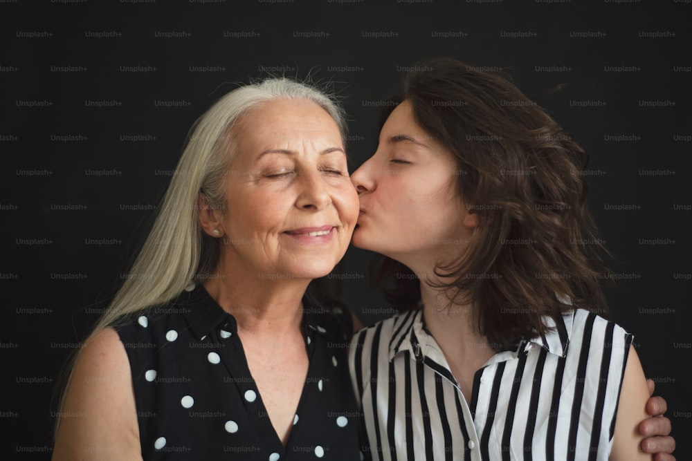 A portrait of senior grandmother with her granddaughter kissing her over black blackground
