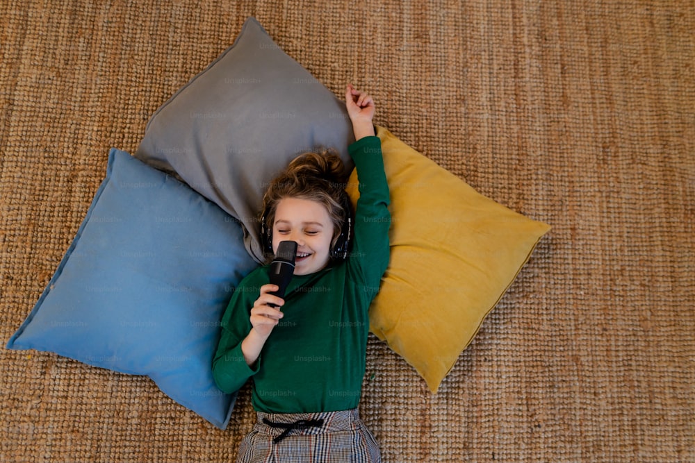 A little girl lying on the floor on cushions listening music,singing having fun and playing at home.