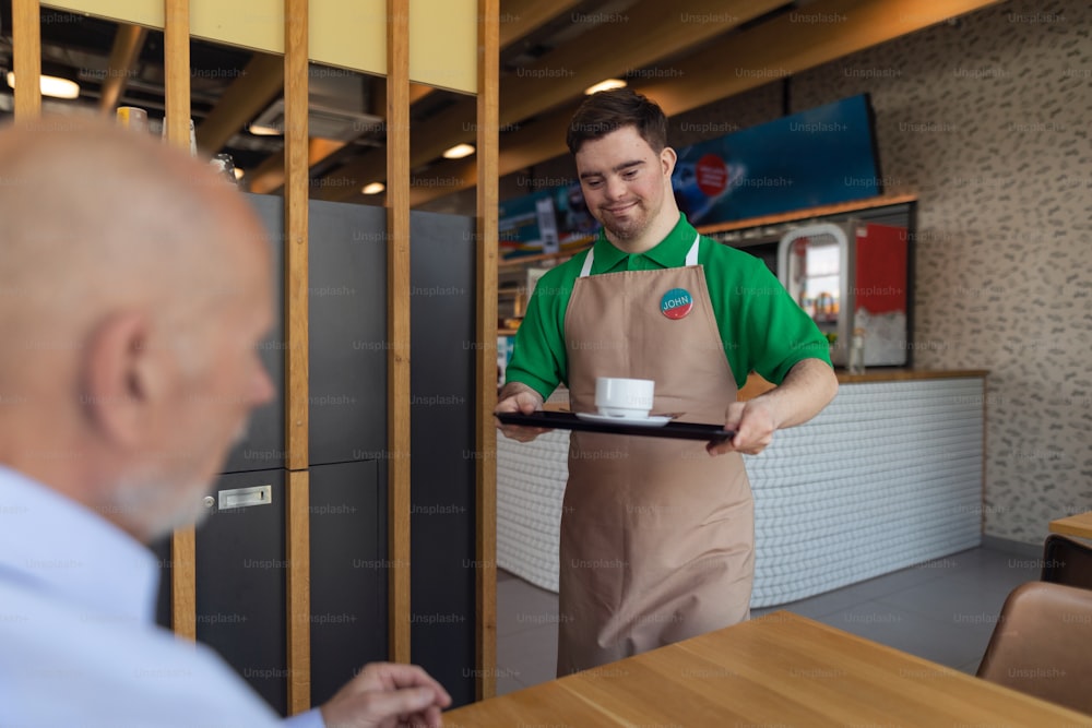 A happy waiter with Down syndrome carrying coffee to customer in cafe at gas station. Social inclusion concept.