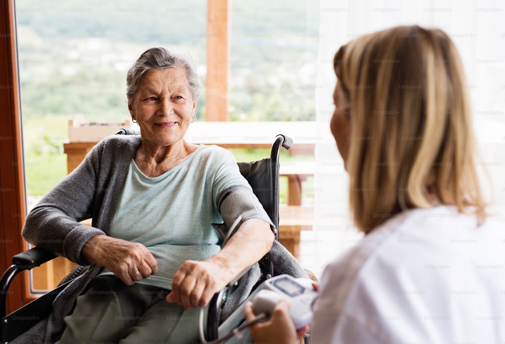 Health visitor and a senior woman during home visit. A nurse checking blood pressure of a woman in an wheelchair.