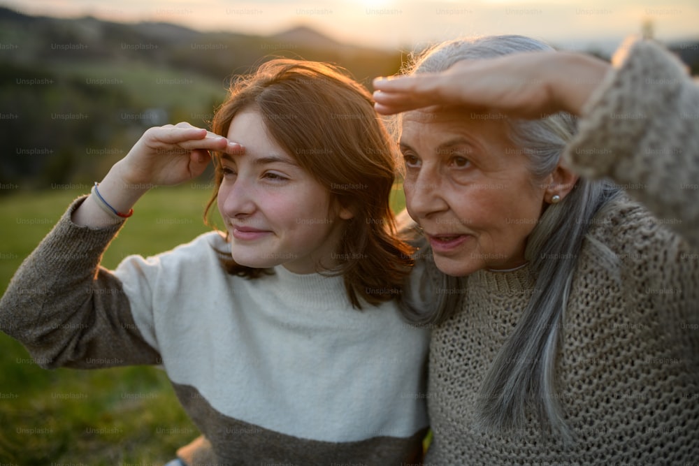 Portrait of a happy senior grandmother with teenage granddaguhter sitting on grass and looking into the distance in nature on spring day during sunset.