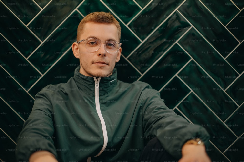 A portrait of young man in jacket and glasses sitting agiant dark tiled wall.
