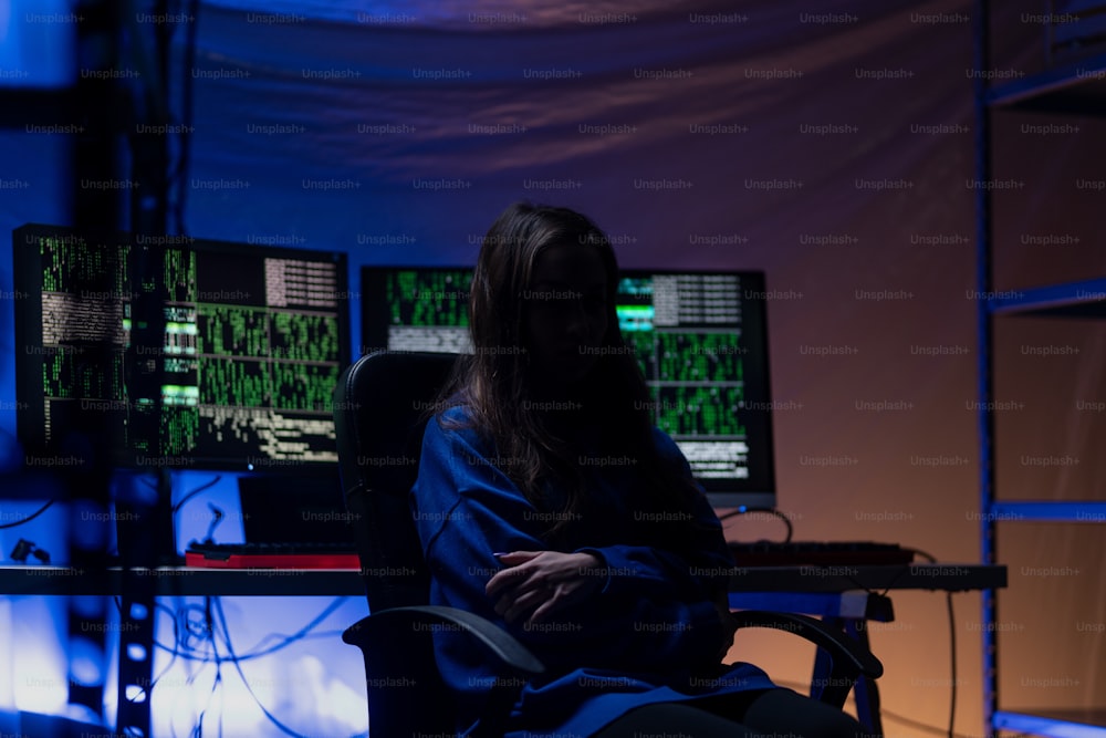 Uma mulher hacker anônima encapuzada pelo computador no quarto escuro à noite, conceito de guerra cibernética.