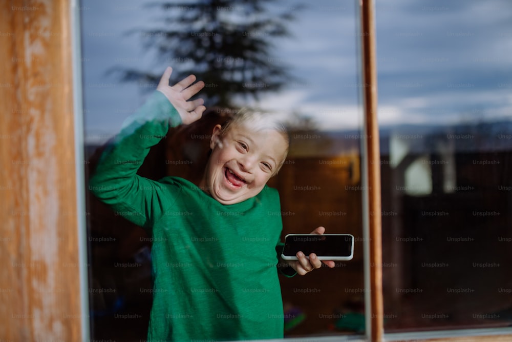 다운 증후군을 앓고 있는 행복한 어린 소년이 스마트폰을 사용하고 창문을 통해 손을 흔든다