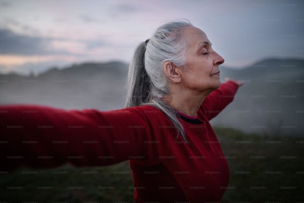 Eine ältere Frau, die am frühen Morgen mit Nebel und Bergen im Hintergrund Atemübungen in der Natur macht.