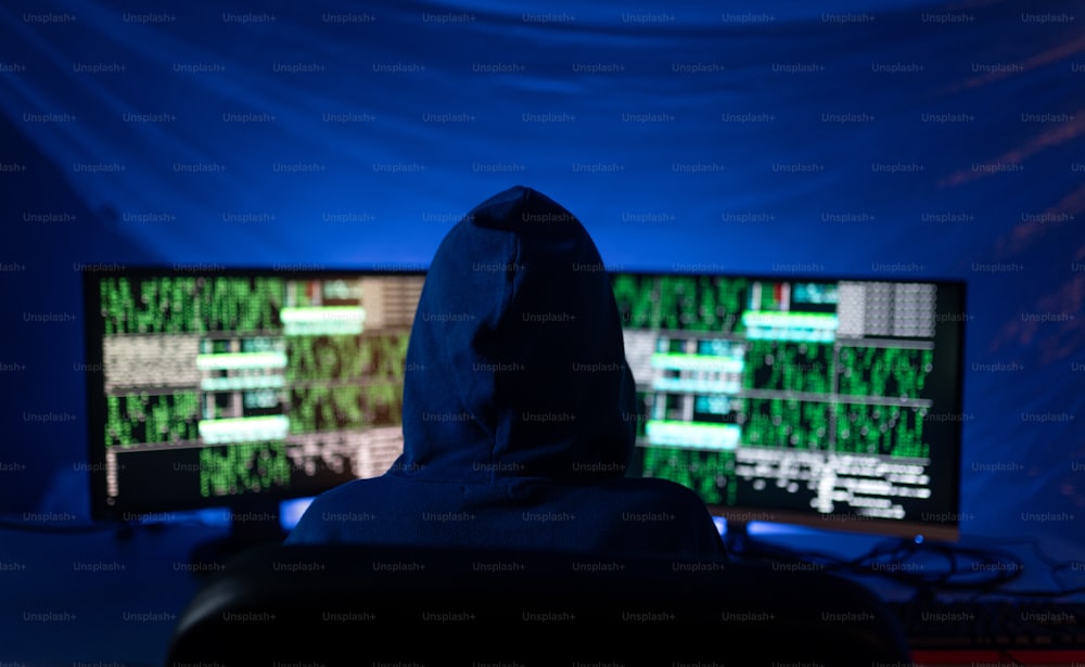 Um hacker com capuz de visão traseira por computador no quarto escuro à noite, conceito de guerra cibernética.