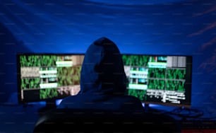 Um hacker com capuz de visão traseira por computador no quarto escuro à noite, conceito de guerra cibernética.