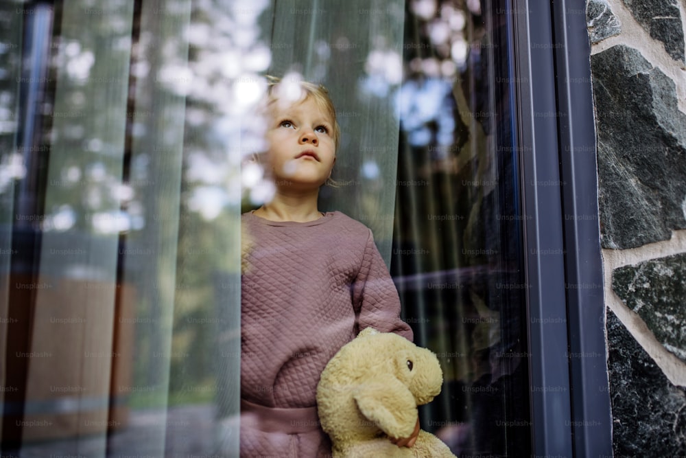 Sad little child standing alone with teddy bear behind the window, photo trough glass.