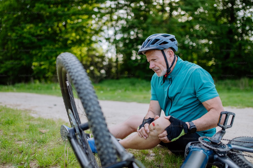 An active senior man in sportswear fell off bicycle on the ground and hurt his knee, in park in summer.