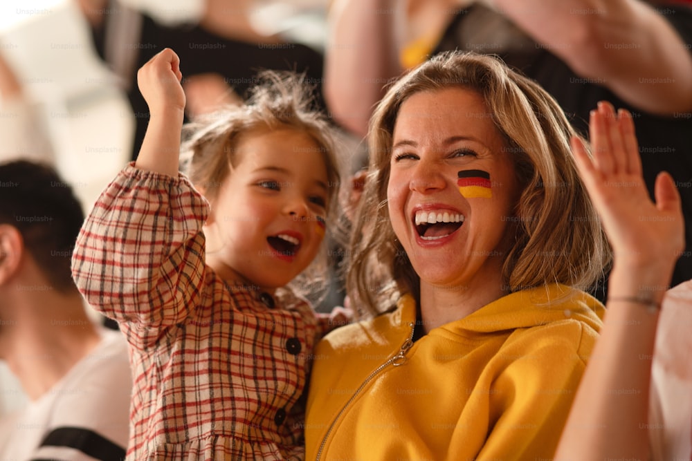 Excited football fans, mother with little daughter, supproting a German national team in live soccer match at stadium.