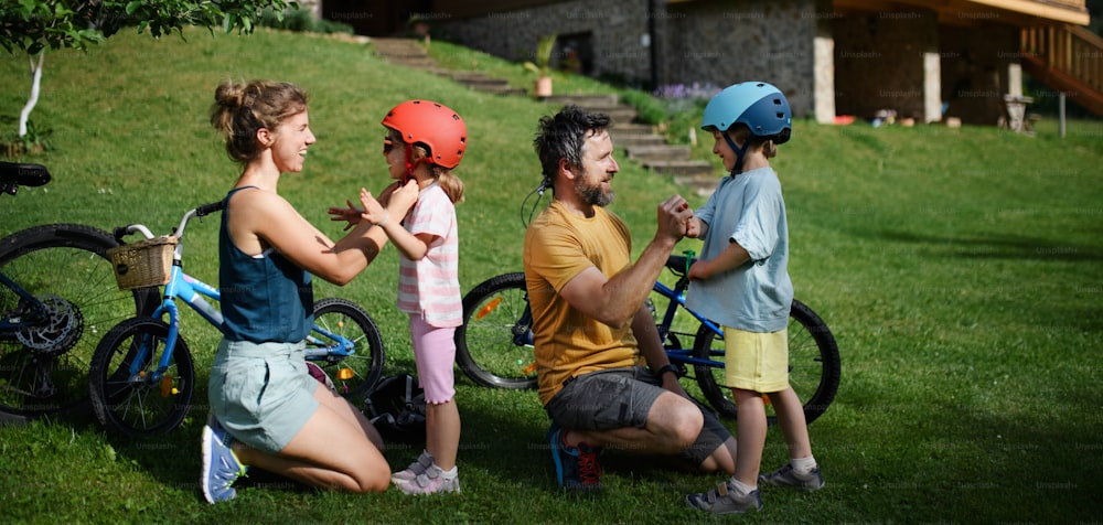 A young family with little children preapring for bike ride, putting on helmets in front of house.