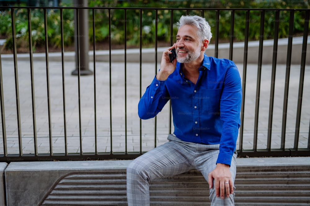 A successful happy businessman waiting in city street, using mobile phone, commuting concept.