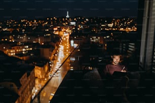 Un joven africano AMerican sentado en un balcón con vista urbana y usando tableta por la noche