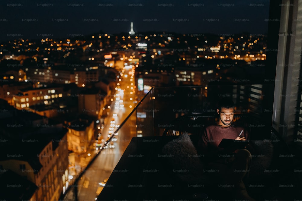 Ein junger afrikanischer Mann sitzt auf einem Balkon mit Blick auf die Stadt und benutzt nachts ein Tablet